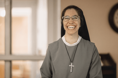 A smiling nun in a gray habit with glasses