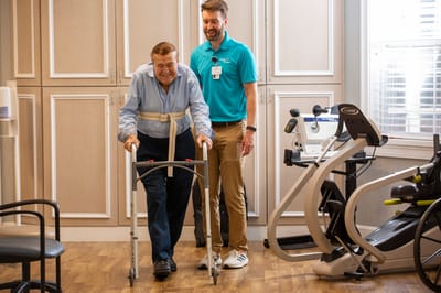 A resident using a walker with staff assistance in a fitness area