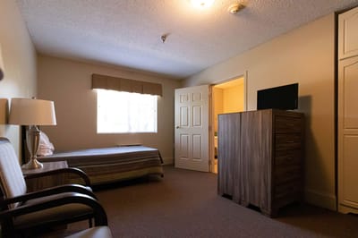 A private resident room with a bed, dresser, and lamp