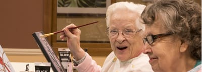 Two senior women engaged in painting activity indoors.