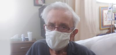 Elderly individual sitting in a room wearing a mask.