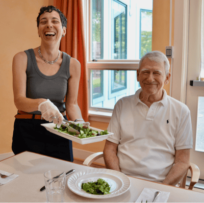 Staff serving salad to a smiling resident in a dining room