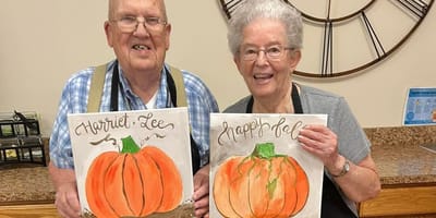 Two residents display their pumpkin paintings in an activity room