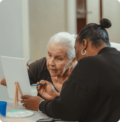An elderly woman painting with a staff member's guidance.
