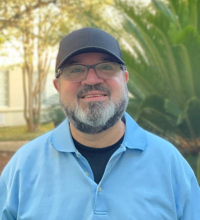 A smiling man outdoors in a blue shirt with greenery behind