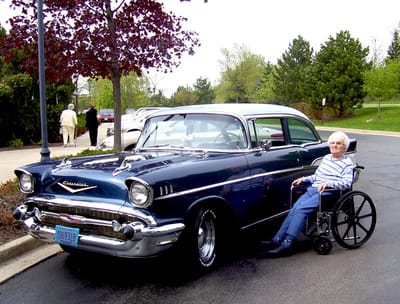 An elderly woman in a wheelchair beside a vintage car