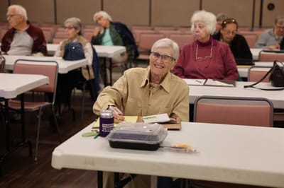 An elderly woman smiling in a common area filled with residents