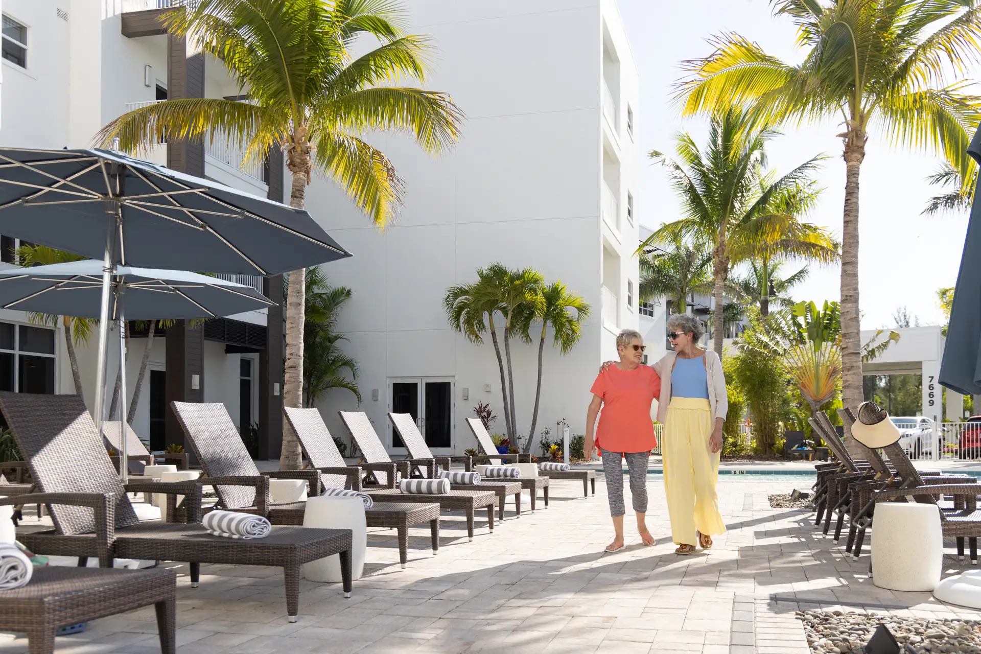 Two women walking by a poolside area with palm trees