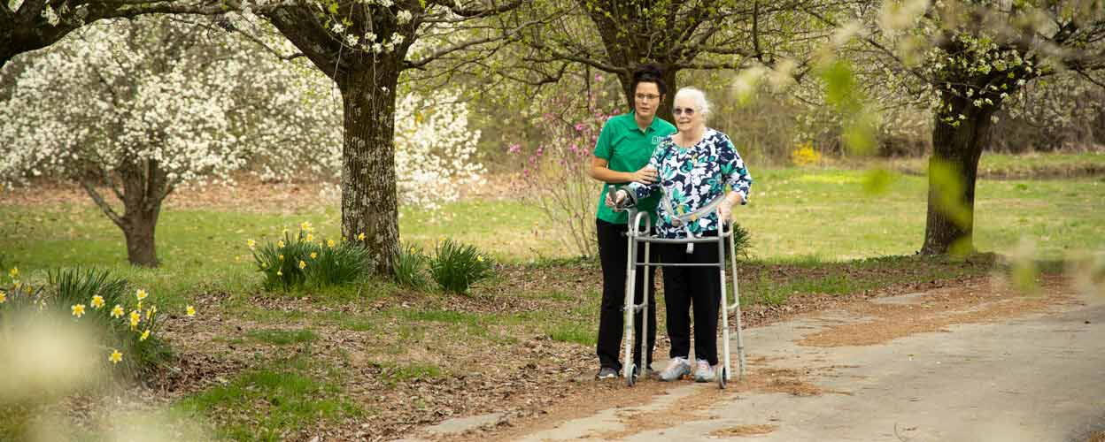 Caregiver helping resident outdoors with a walker.