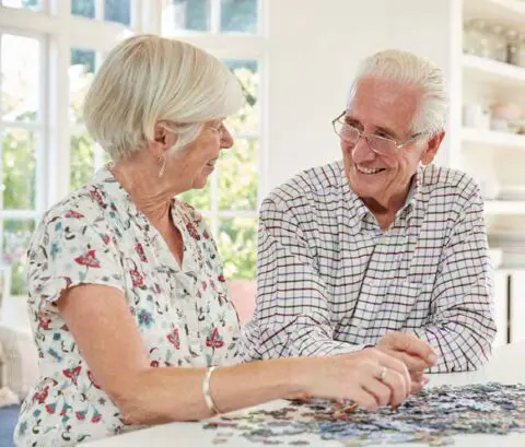 Two seniors smiling and doing a puzzle indoors
