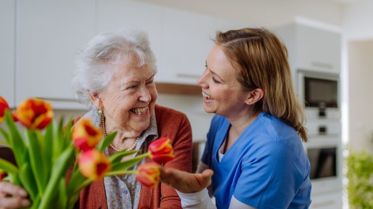Senior resident smiling with caregiver and flowers.