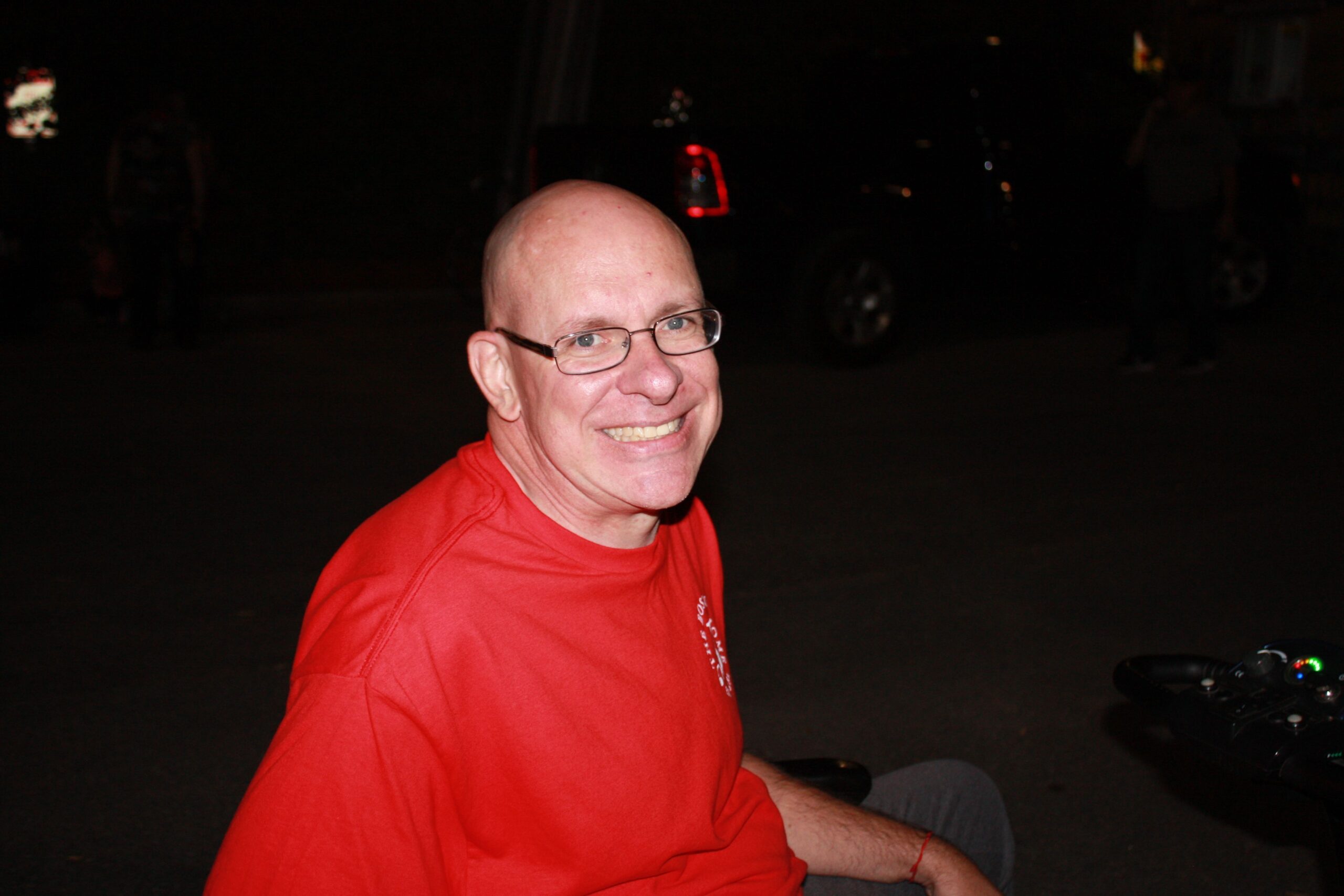 Man smiling while seated outdoors in a red shirt.