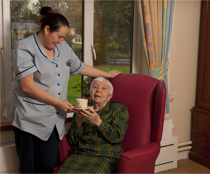 Staff member serving tea to a resident in a cozy chair.