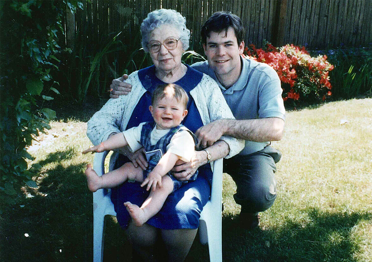 An elderly woman holding a baby with a young man outdoors.
