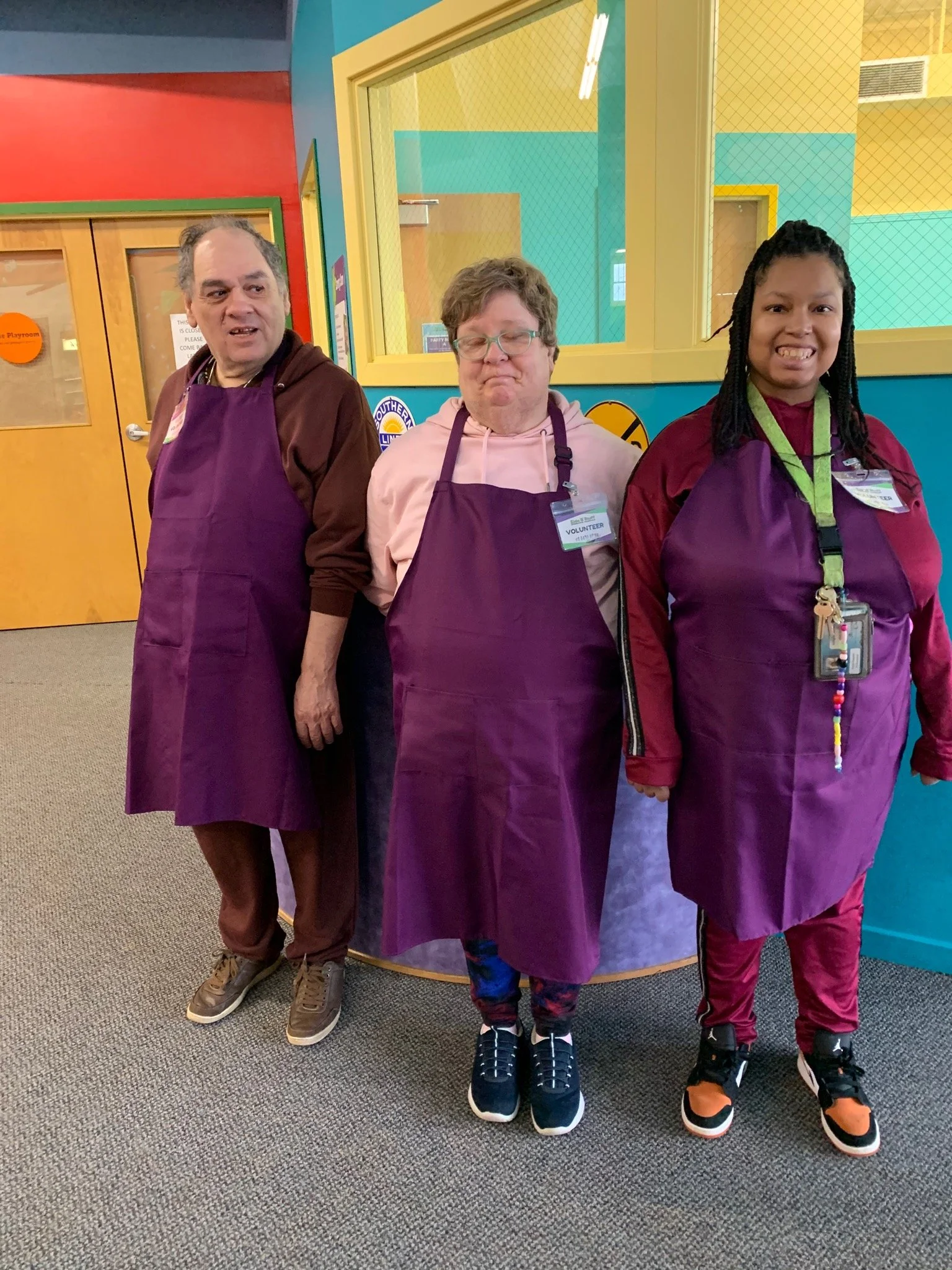Three staff members wearing purple aprons in a colorful common area.