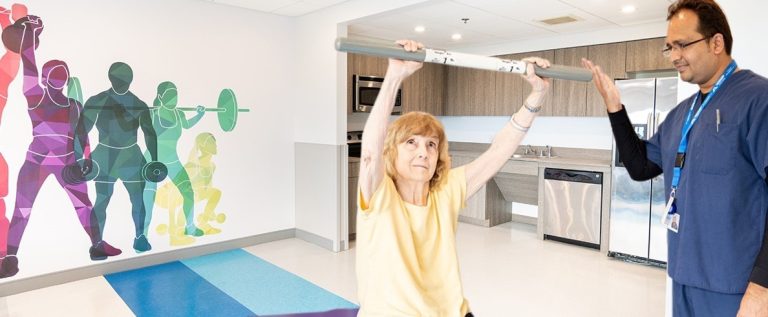 An elderly woman exercising with staff support.