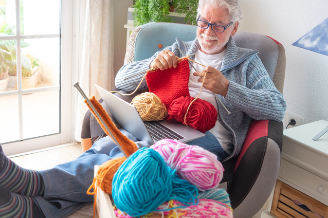 Senior man knitting with yarn and laptop.