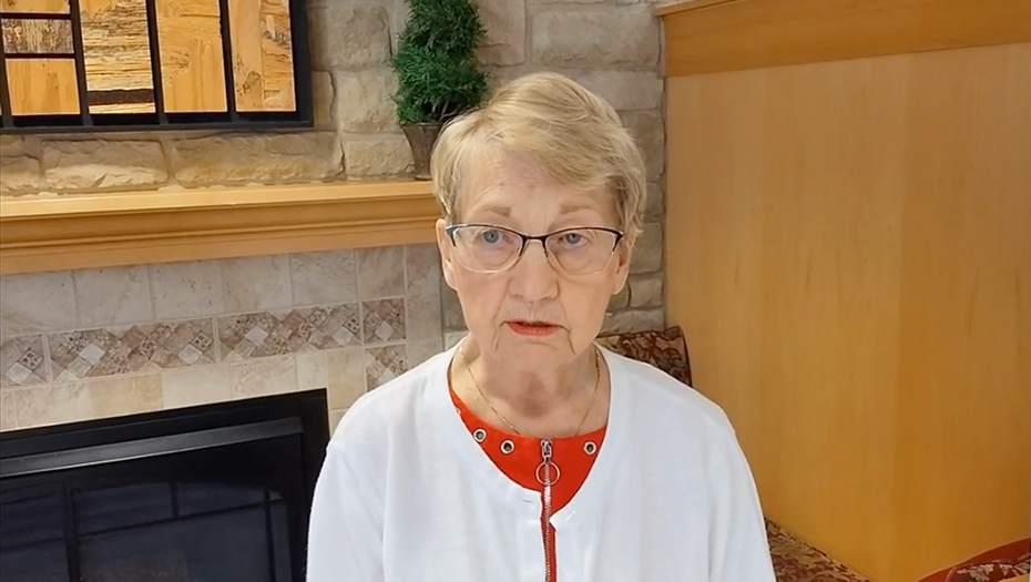 An elderly woman speaking indoors near a fireplace