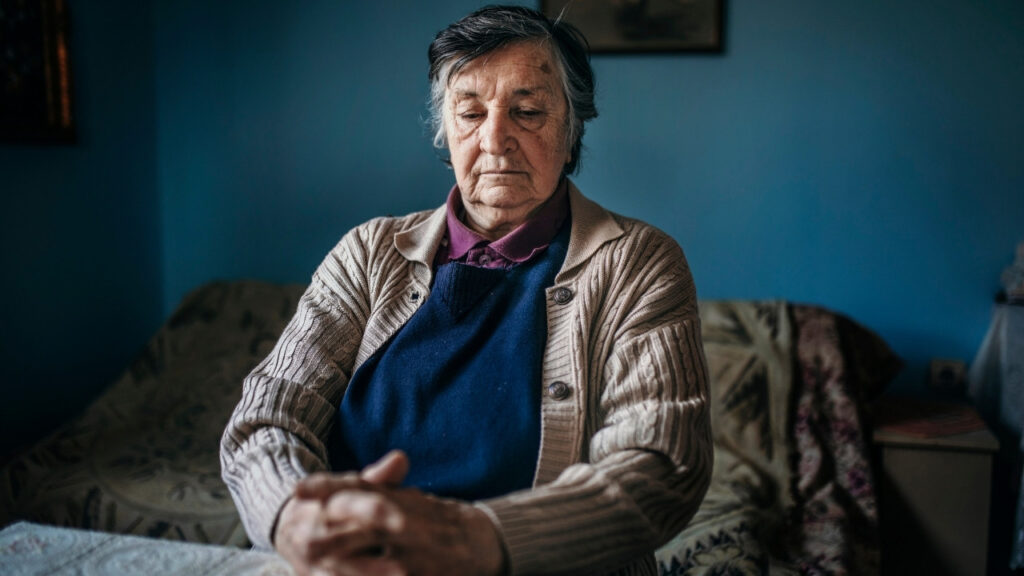 An elderly woman seated quietly indoors, reflecting.
