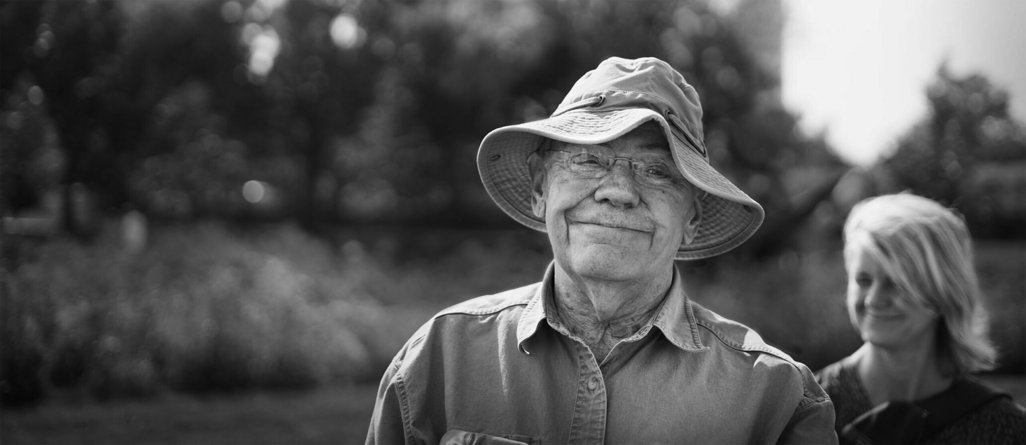Smiling elderly man outdoors with a woman in the background.