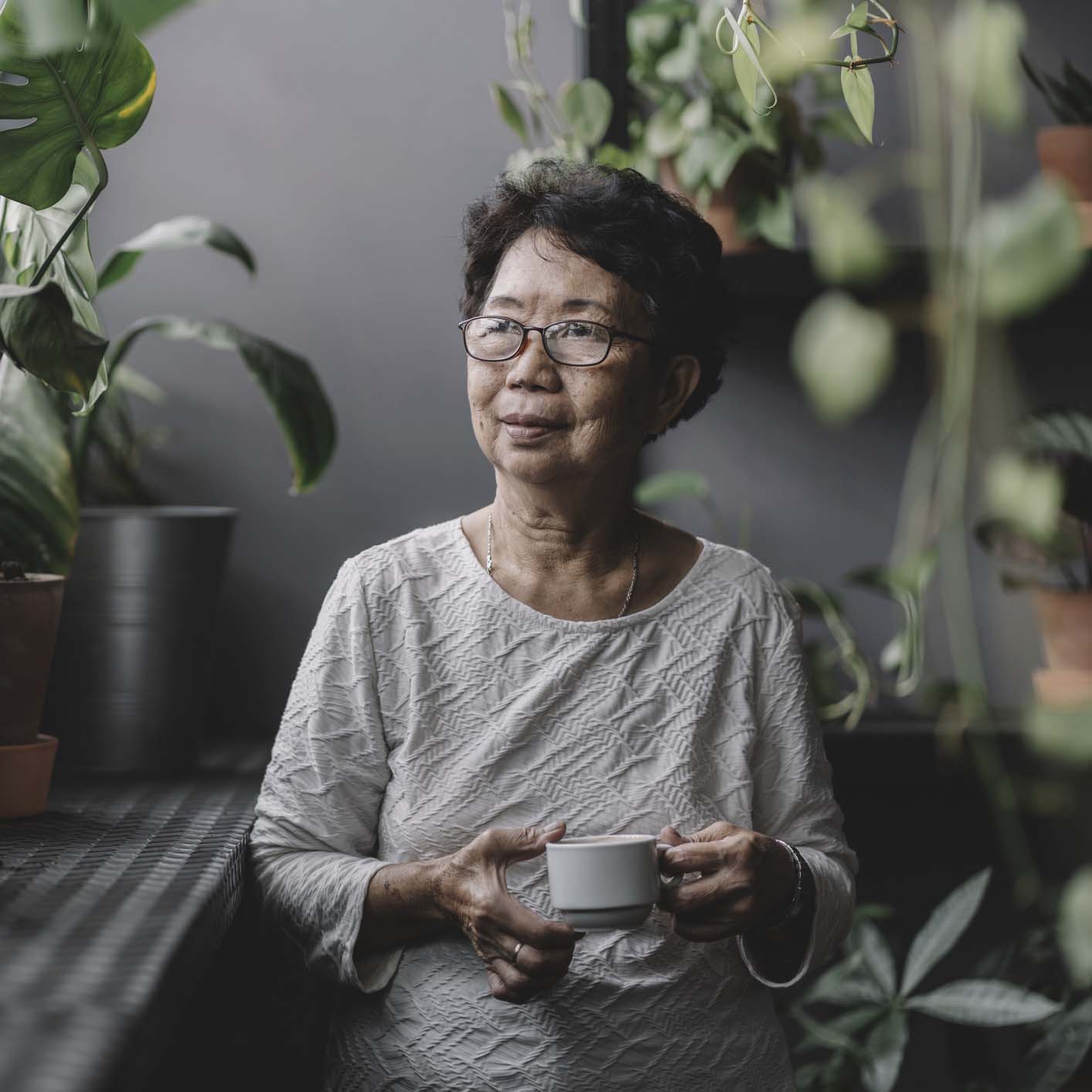Senior woman enjoying a cup in a plant-filled interior.