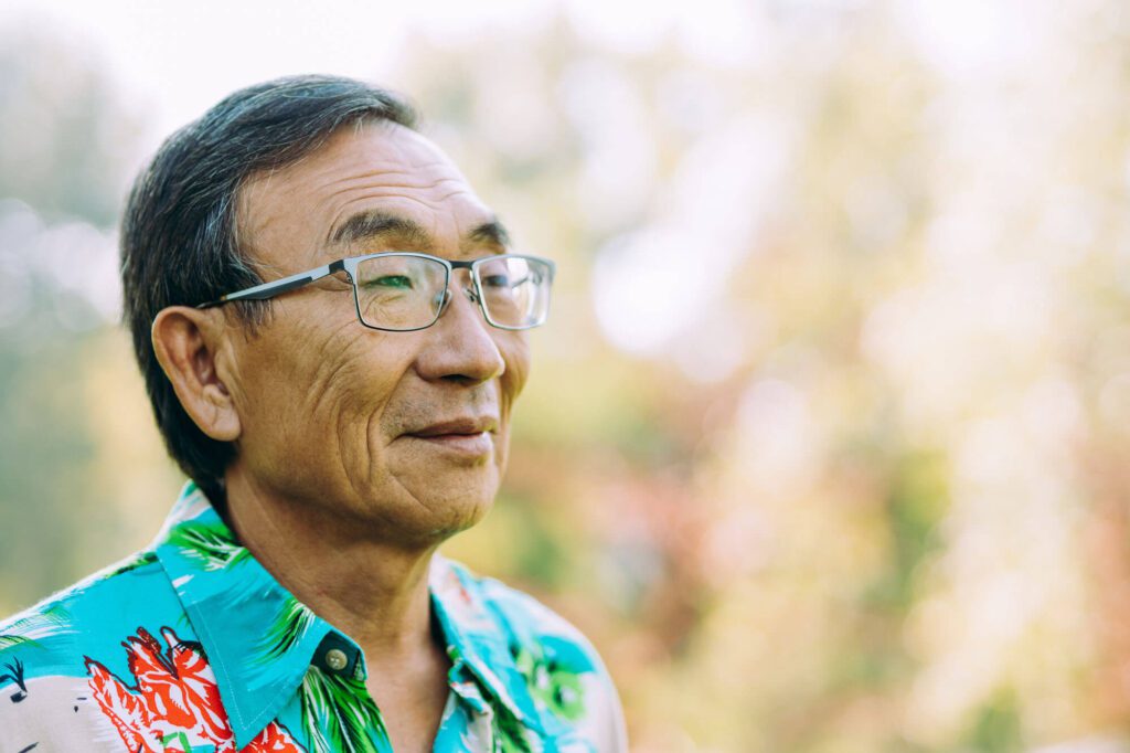 Elderly man smiling outdoors wearing a floral shirt.
