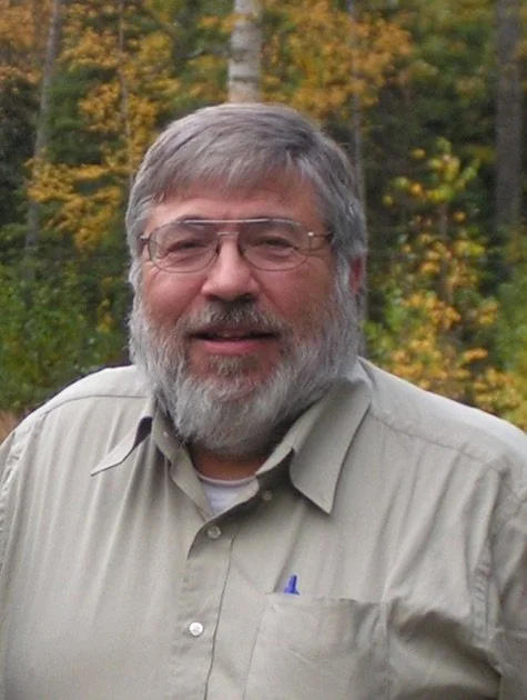 A smiling man outdoors with a forest background.