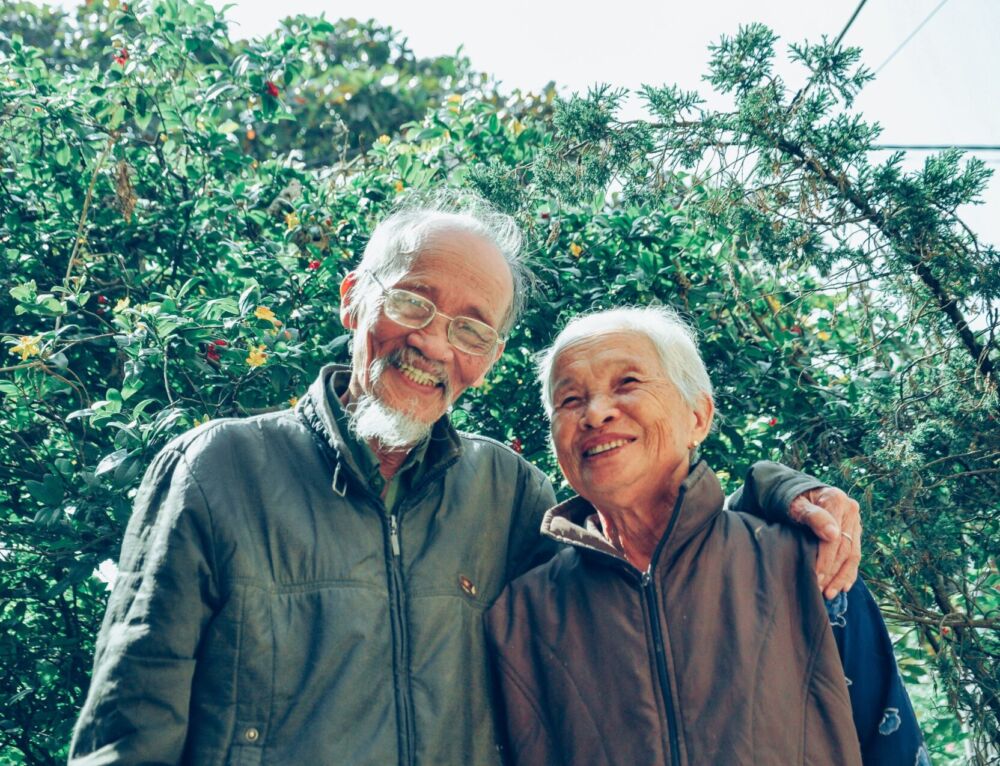 Two elderly individuals smiling outdoors in a garden.