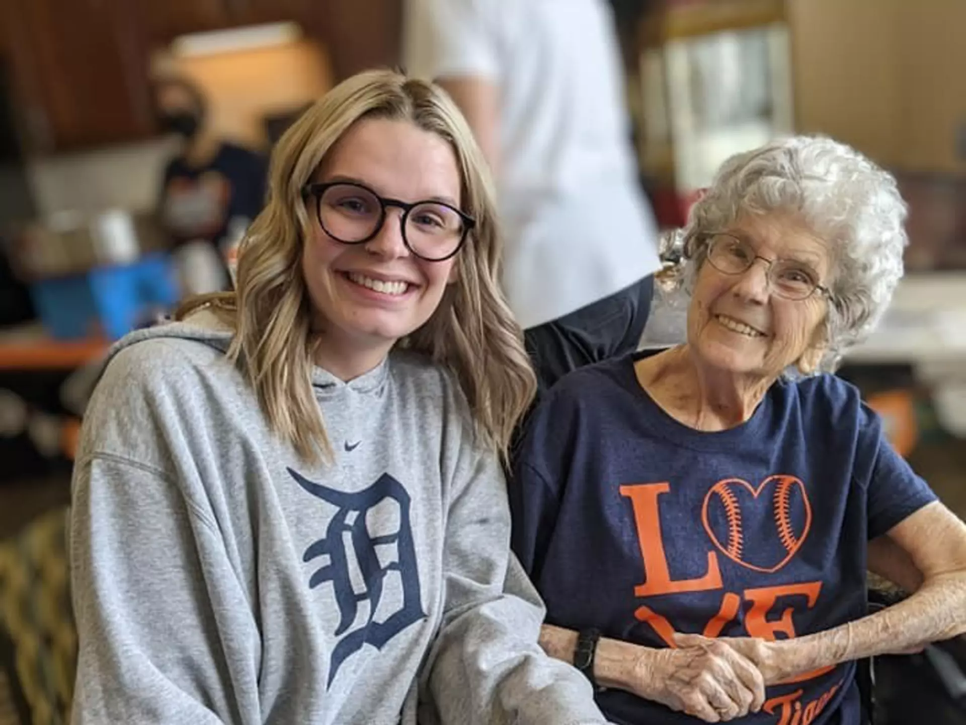 A woman with glasses smiling alongside an elderly lady in a common area