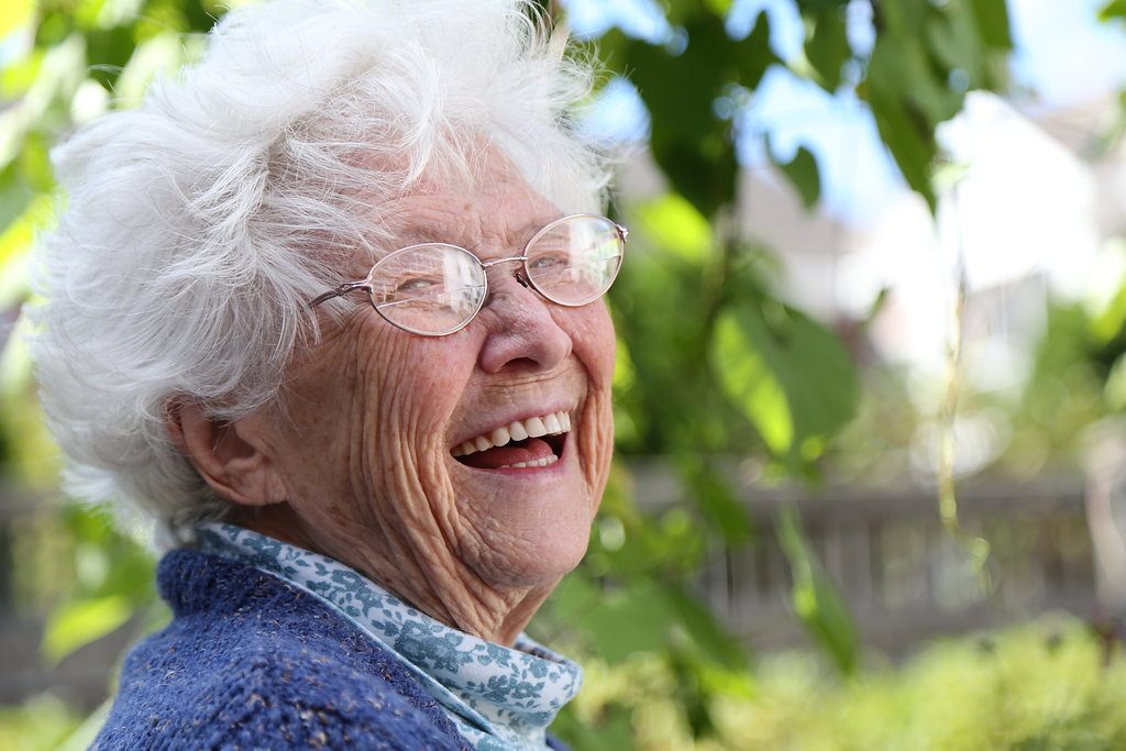 Elderly woman smiling outdoors with greenery in background