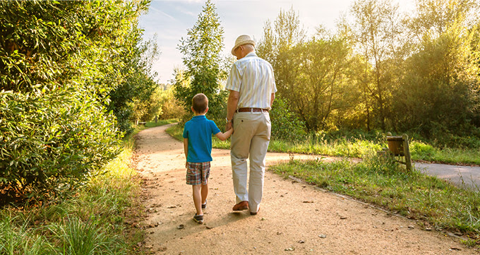 An elderly man walking with a child on a sunny path.