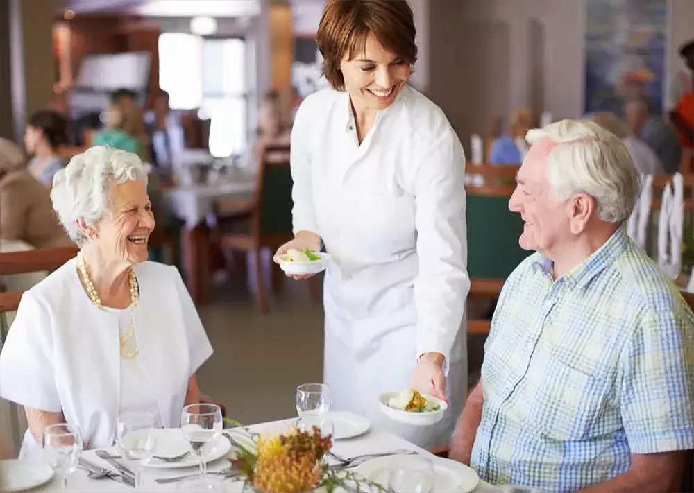 A caregiver serving a meal to smiling residents in the dining room