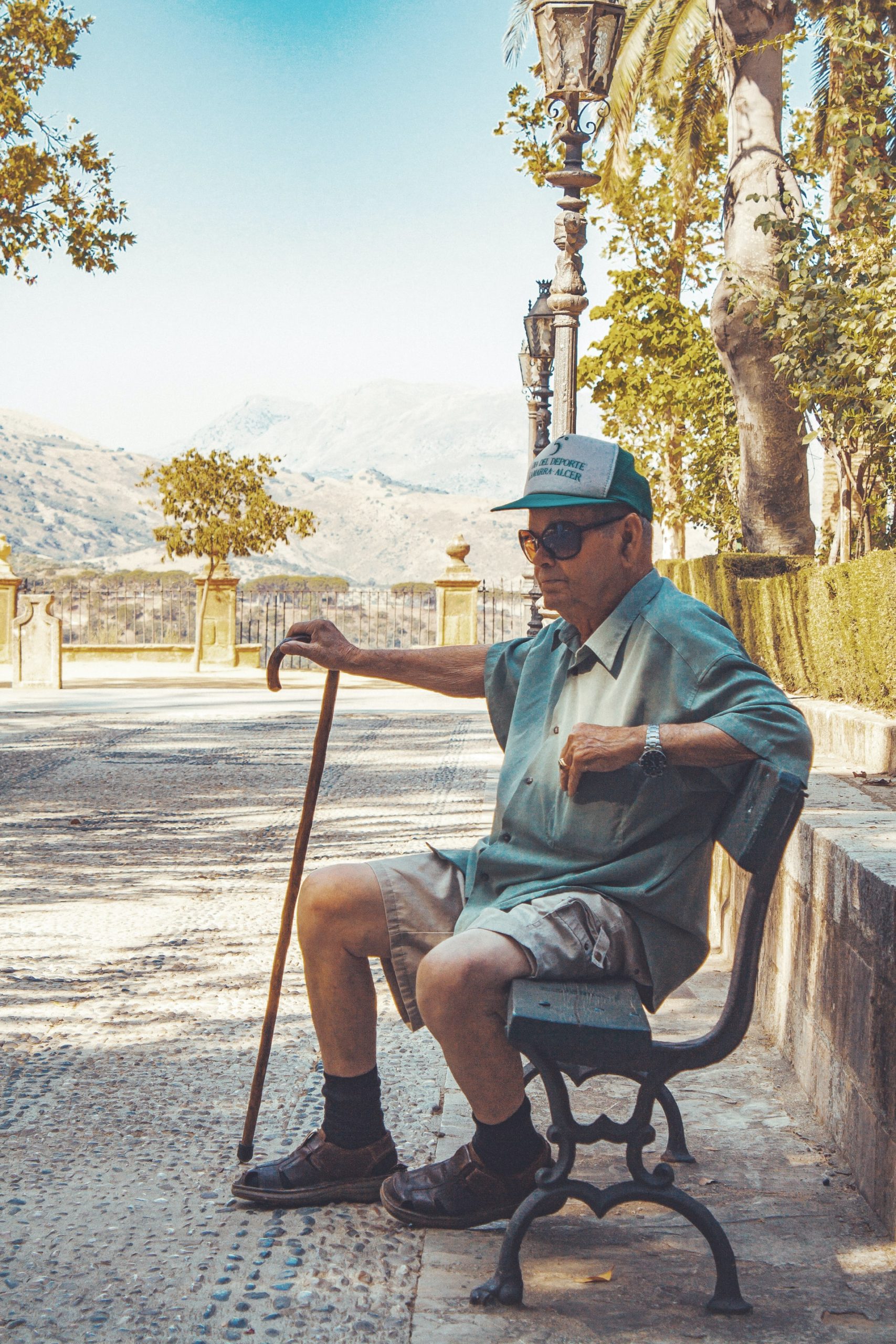 An elderly man sitting on a bench outdoors with a cane.