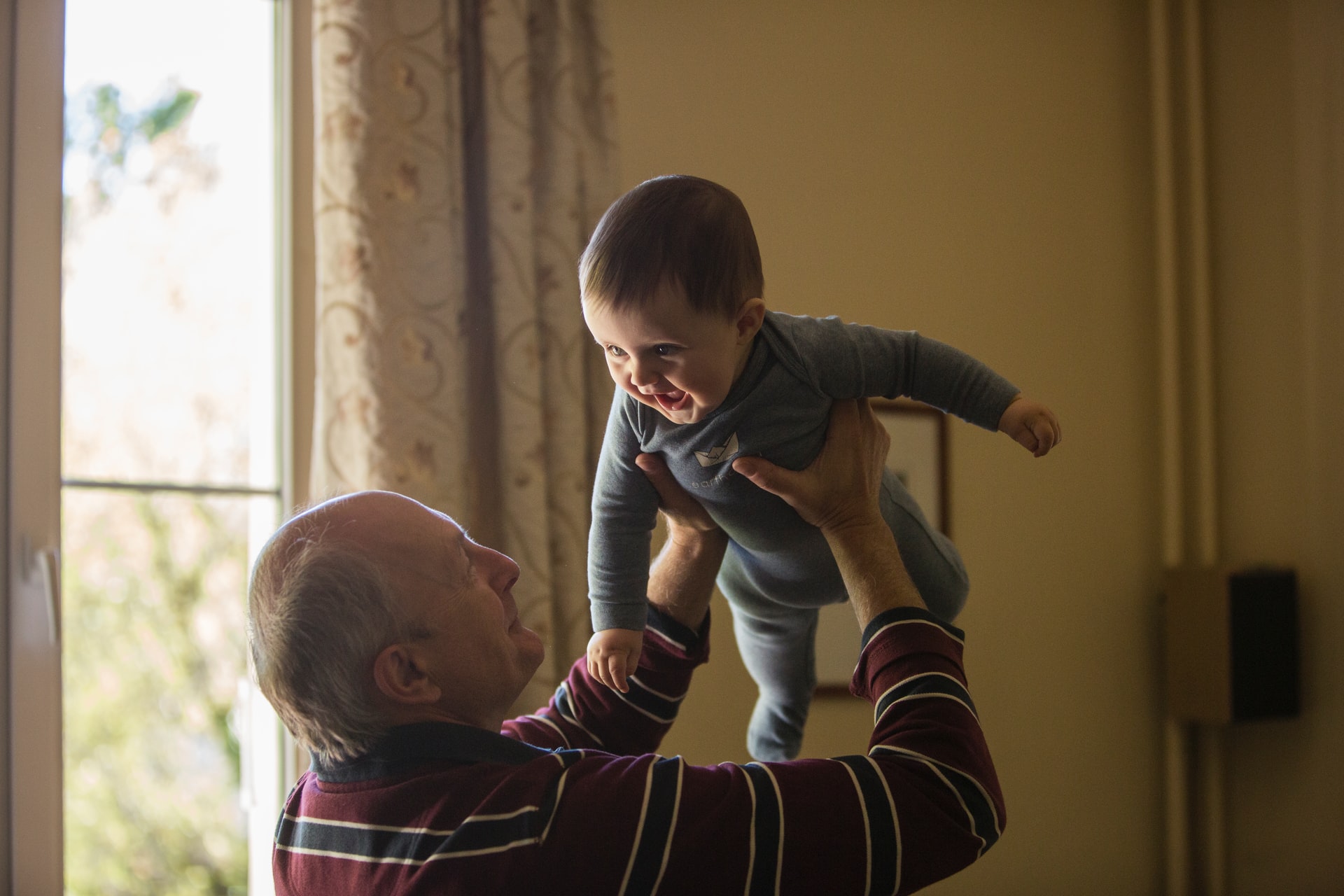 A senior man joyfully holds a baby indoors.