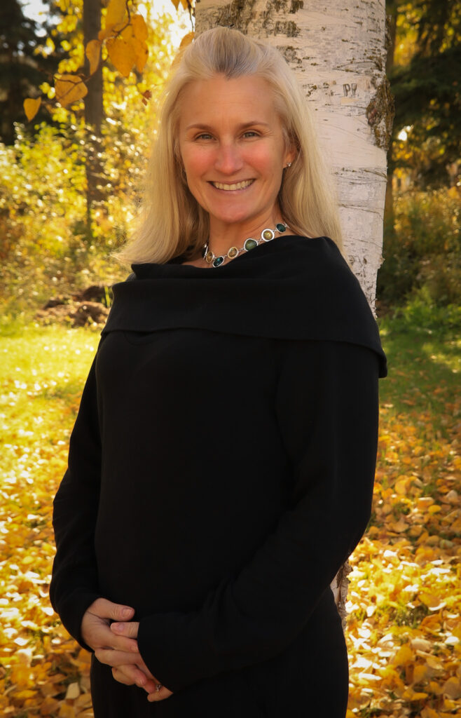Woman smiling in an outdoor setting with fall foliage.