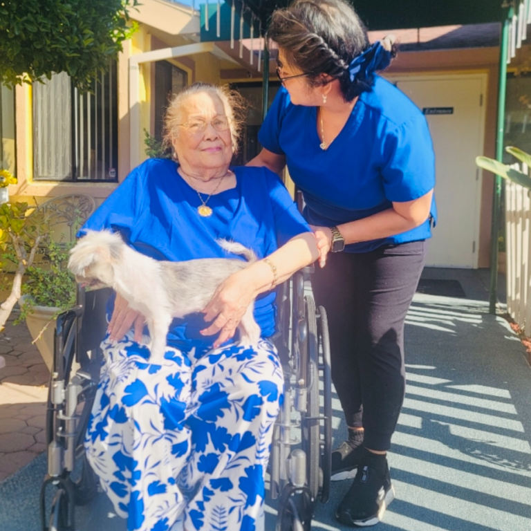 Resident in wheelchair with staff and dog outdoors.