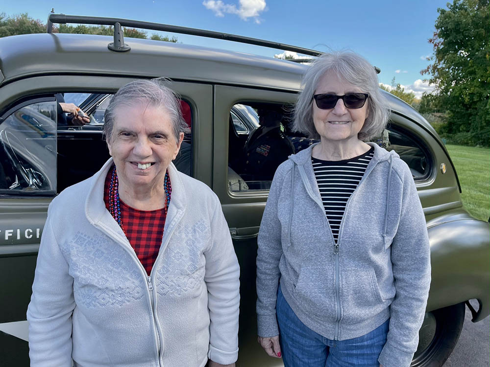 Two residents smiling outside near a vintage vehicle