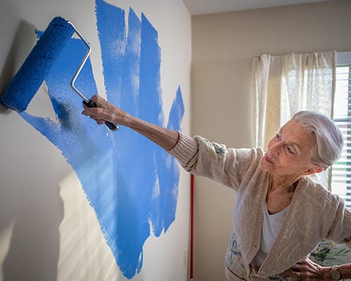 An elderly woman painting a wall blue in a bright room