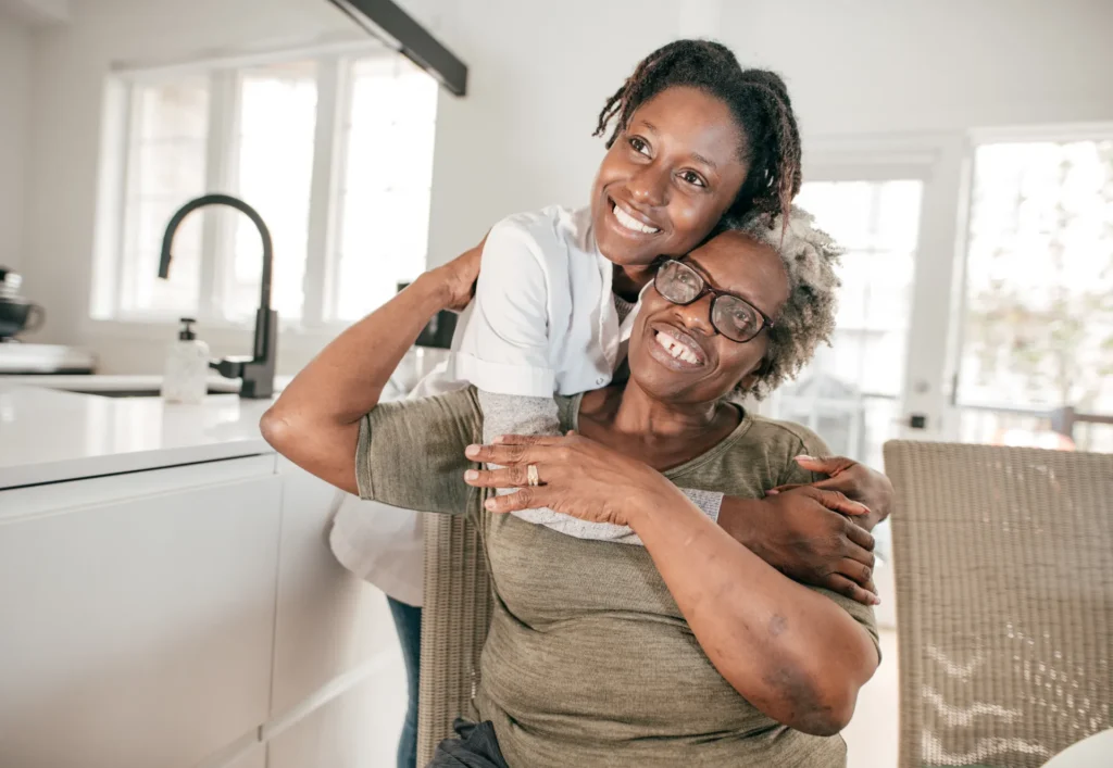 A caregiver hugging a smiling elderly woman indoors.