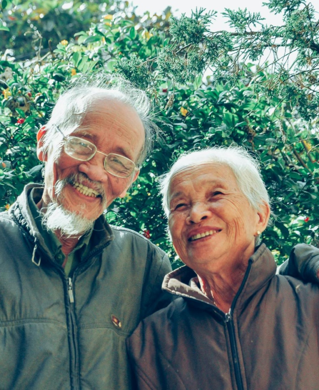Elderly couple smiling outdoors among greenery.