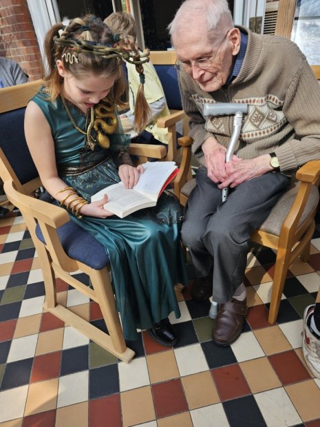 A young girl reading to an elderly man in a cozy setting.
