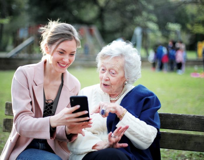 A caregiver shows a smartphone to a senior woman outdoors.