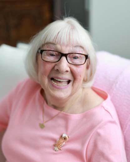 A smiling elderly woman in a pink shirt with glasses