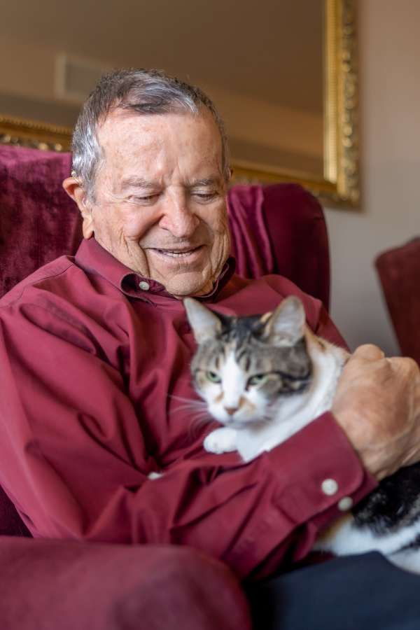 An elderly man smiling while holding a cat in a cozy chair