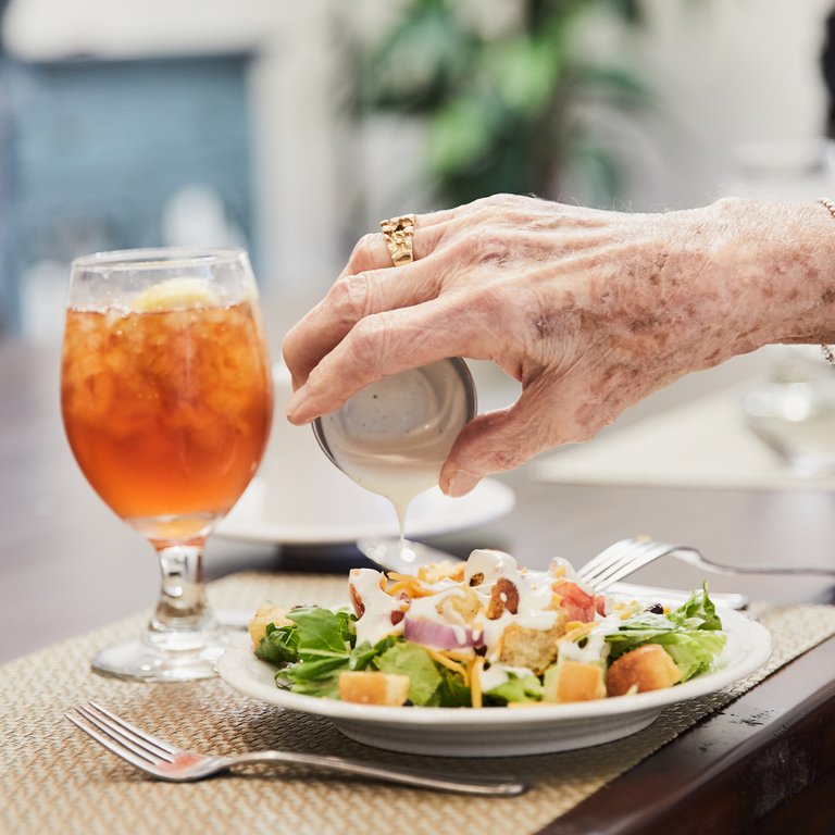 A resident's hand pouring dressing on a salad at lunch