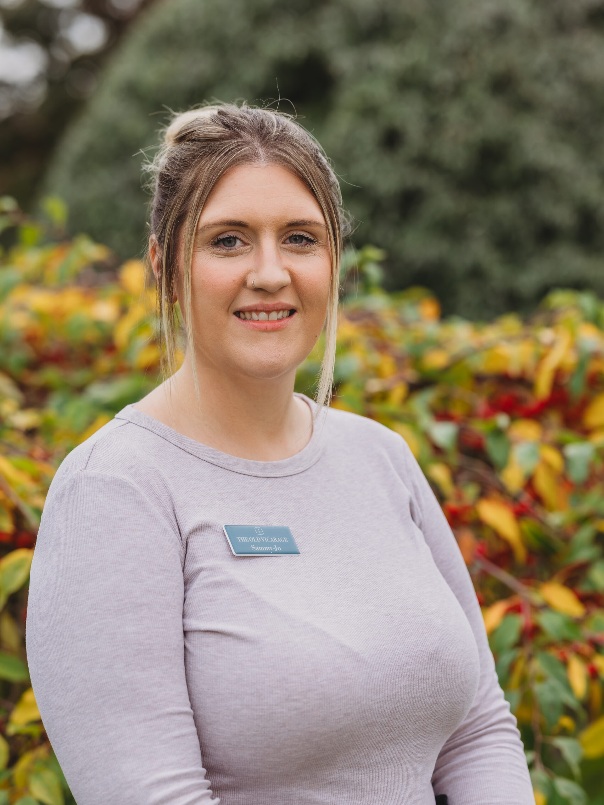 Staff member smiling in an outdoor setting with foliage.