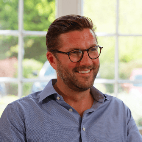 Man smiling indoors with large windows behind.
