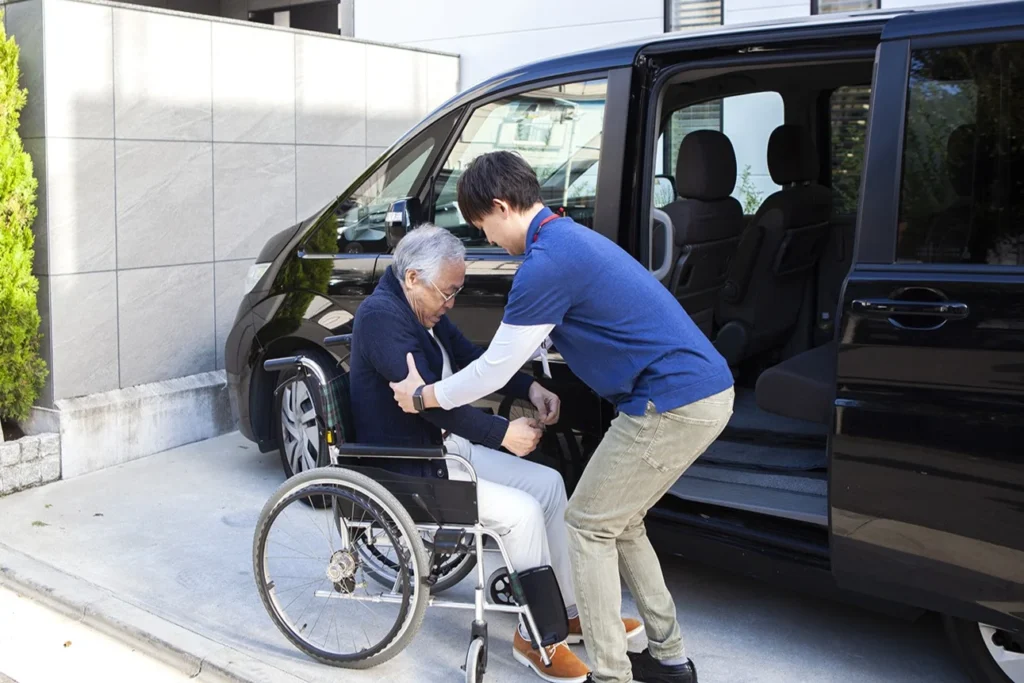 Caregiver assisting a resident in a wheelchair outside a vehicle.