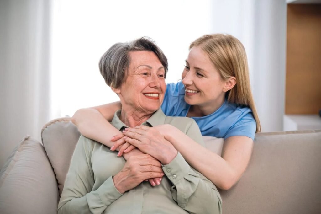 Elderly woman and caregiver smiling together on a couch.
