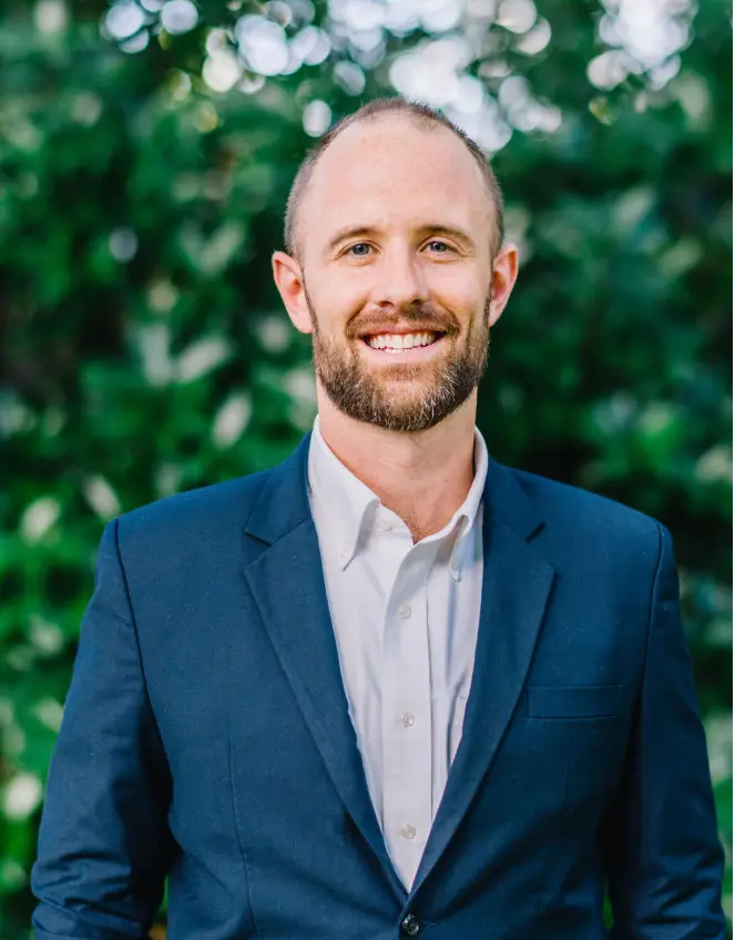 Smiling man in a suit outdoors with greenery.